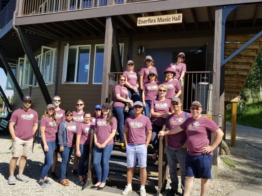 Group of people in matching shirts posing outside the Enerflex Music Hall on a sunny day at Kindle Kids Cancer's Camp Kindle.