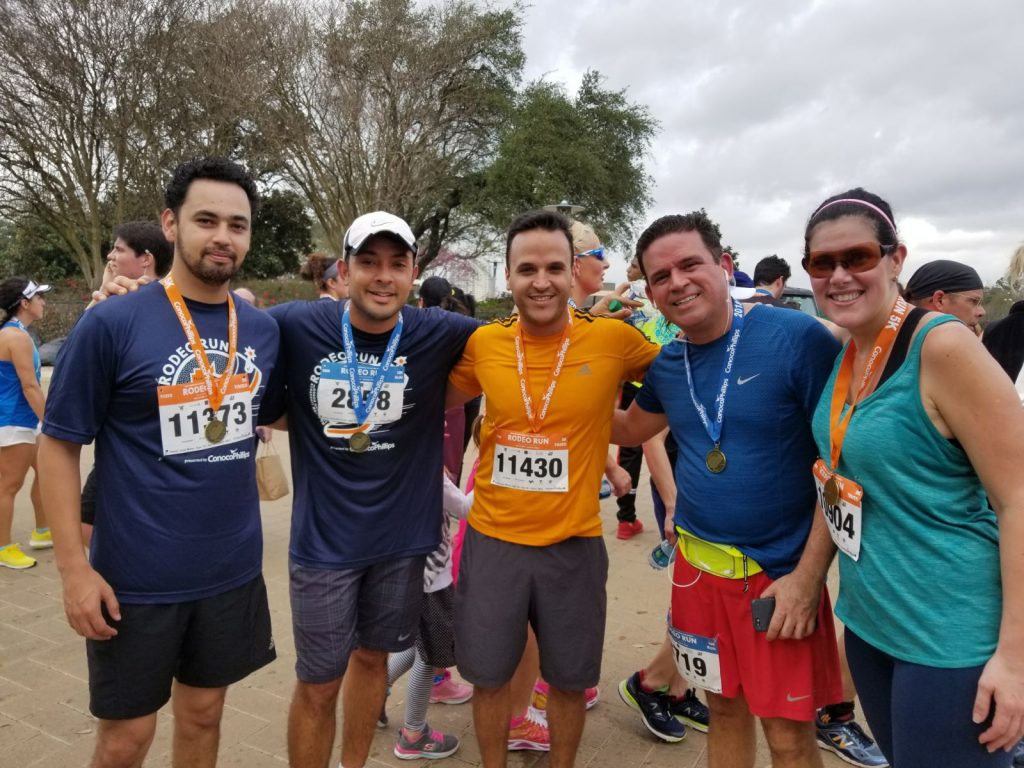 Group of marathon runners wearing medals and bibs, smiling after a race in a park, with trees and other participants in the background.