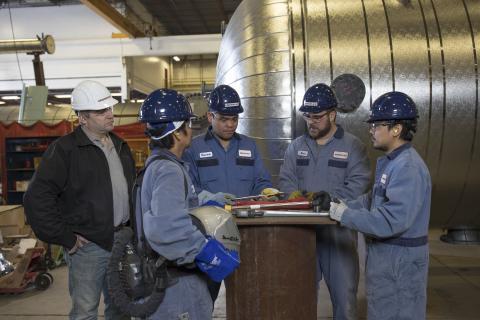 Factory workers in blue uniforms and hard hats having a discussion with a supervisor in a large industrial setting.