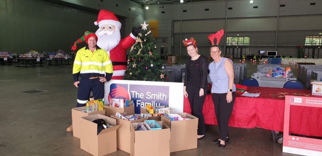 Three people in festive attire stand by donation boxes and a Christmas tree at a holiday charity event by The Smith Family.