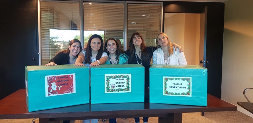 Group of women smiling behind three large wrapped gift boxes on a table, labeled Familia Martinez, Familia Carriso, and Familia Nieve Cortese.