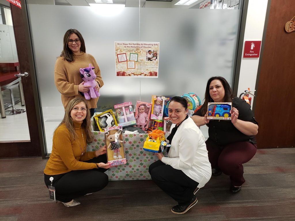 Four women display a variety of toys in front of a gift wrapping station during a holiday charity event.