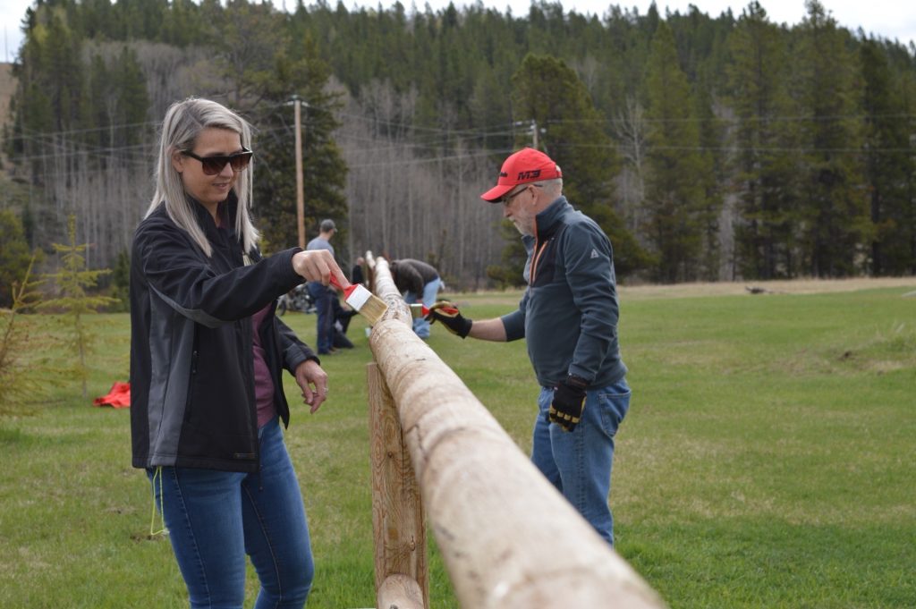 Volunteers painting a wooden fence in a scenic outdoor area with lush greenery and forest background.