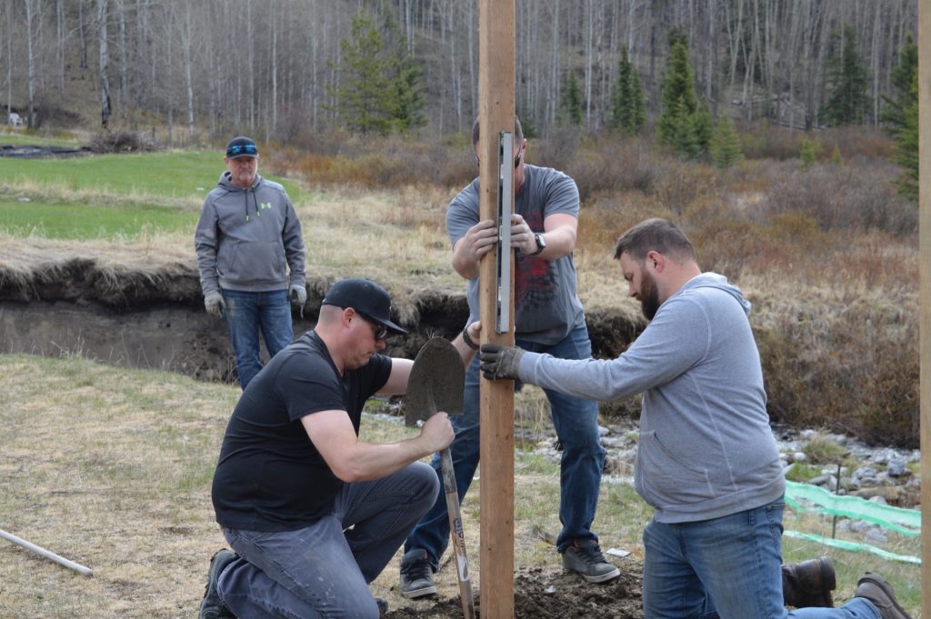 Group of men installing a wooden post in a grassy field with shovels and tools, ensuring it's level.