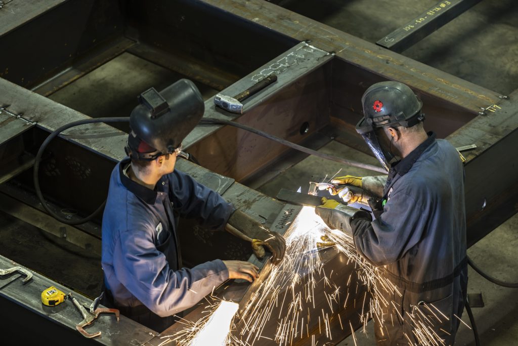 Two industrial workers welding a large metal structure wearing safety gear and generating bright sparks in a factory environment.