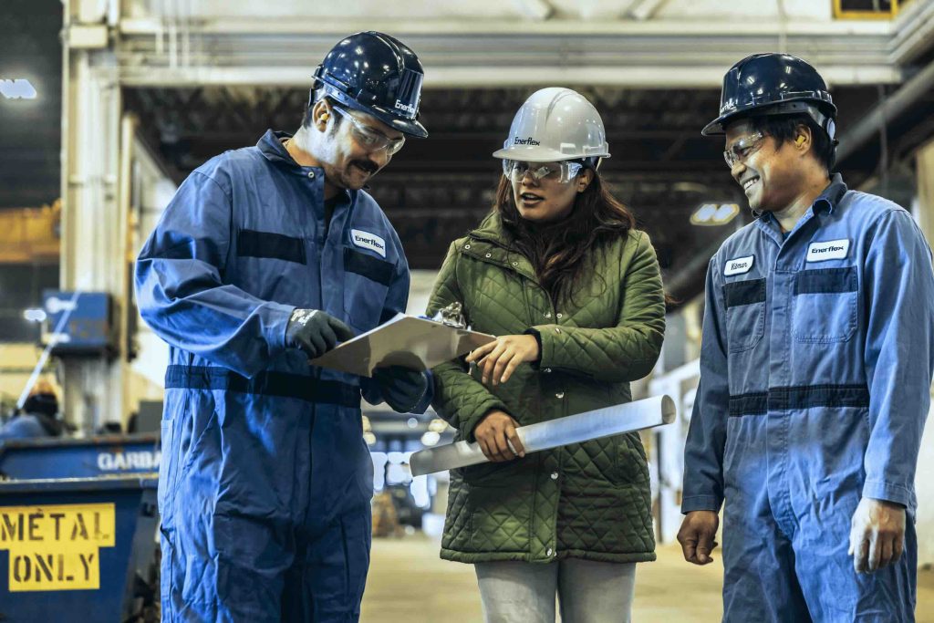 Three Enerflex employees wearing hard hats look over a clipboard and discuss.