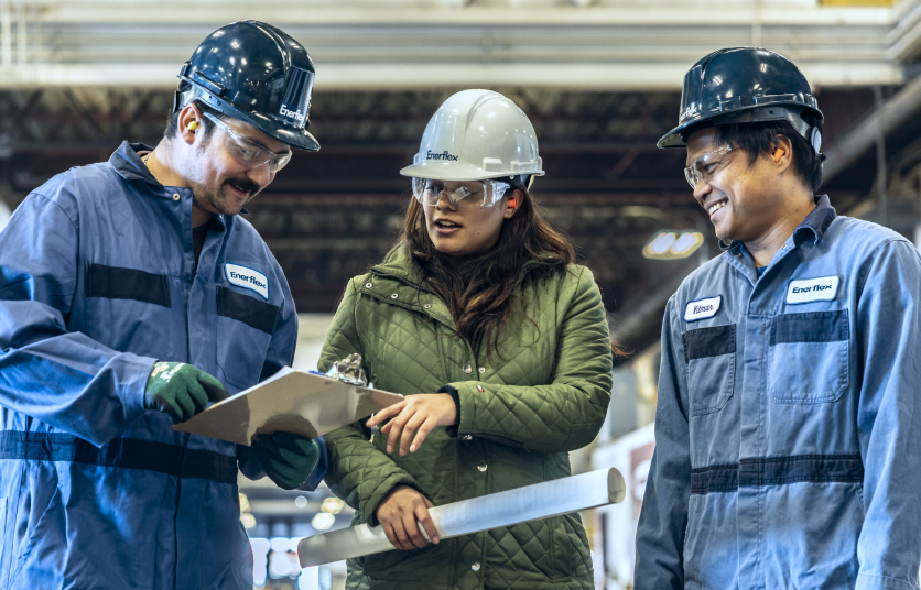 Three Enerflex employees wearing hard hats look over a clipboard and discuss.