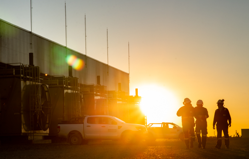 Three Enerflex employees wearing hardhands outside a facility at sunrise. The sun is rising directly behind them, casting bright light over the scene.